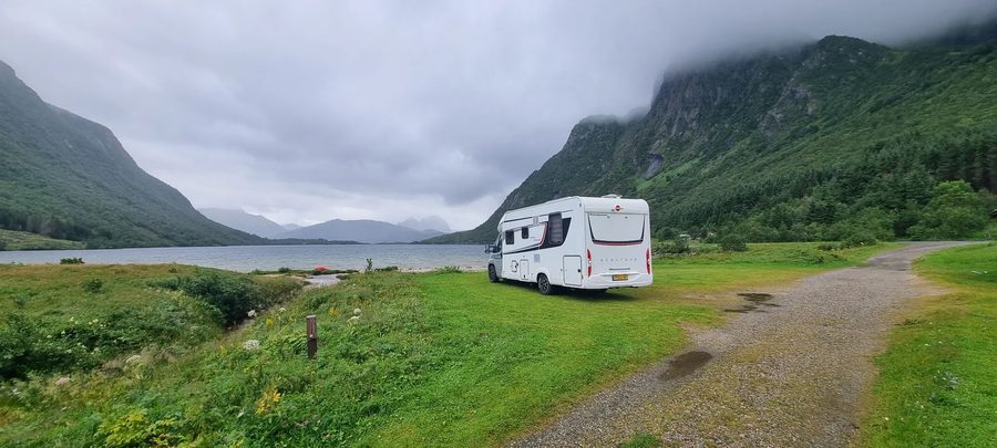 De camper op een groen veld langs een fjord met besneeuwde bergen op de achtergrond